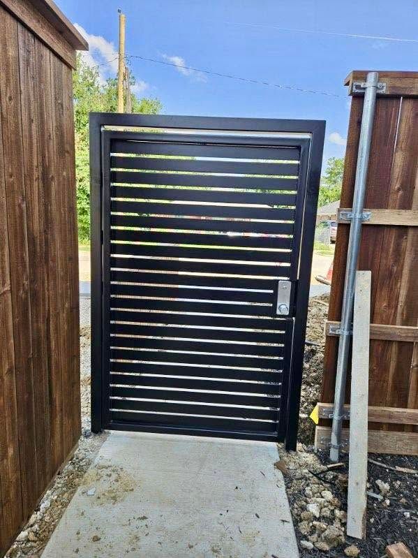 Black slatted gate between wooden fences opening to a narrow concrete path.