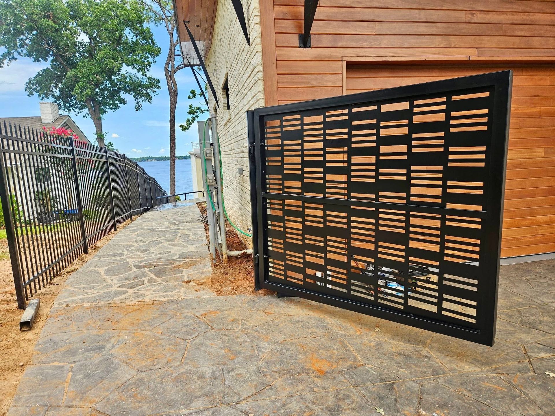 Black metal gate beside a sidewalk next to a building, with trees and water in the distance.
