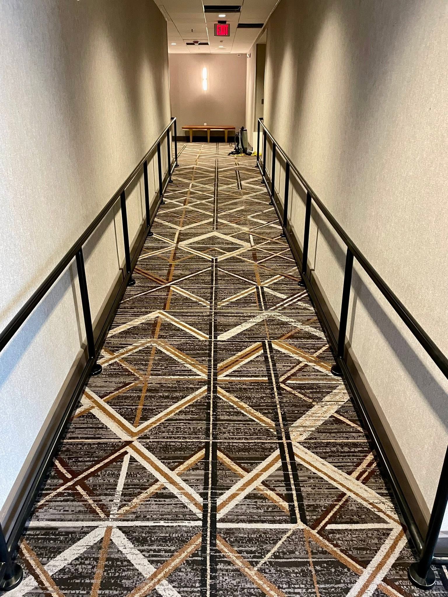 Carpeted hotel hallway with black handrails on both sides and a red exit sign at the end.