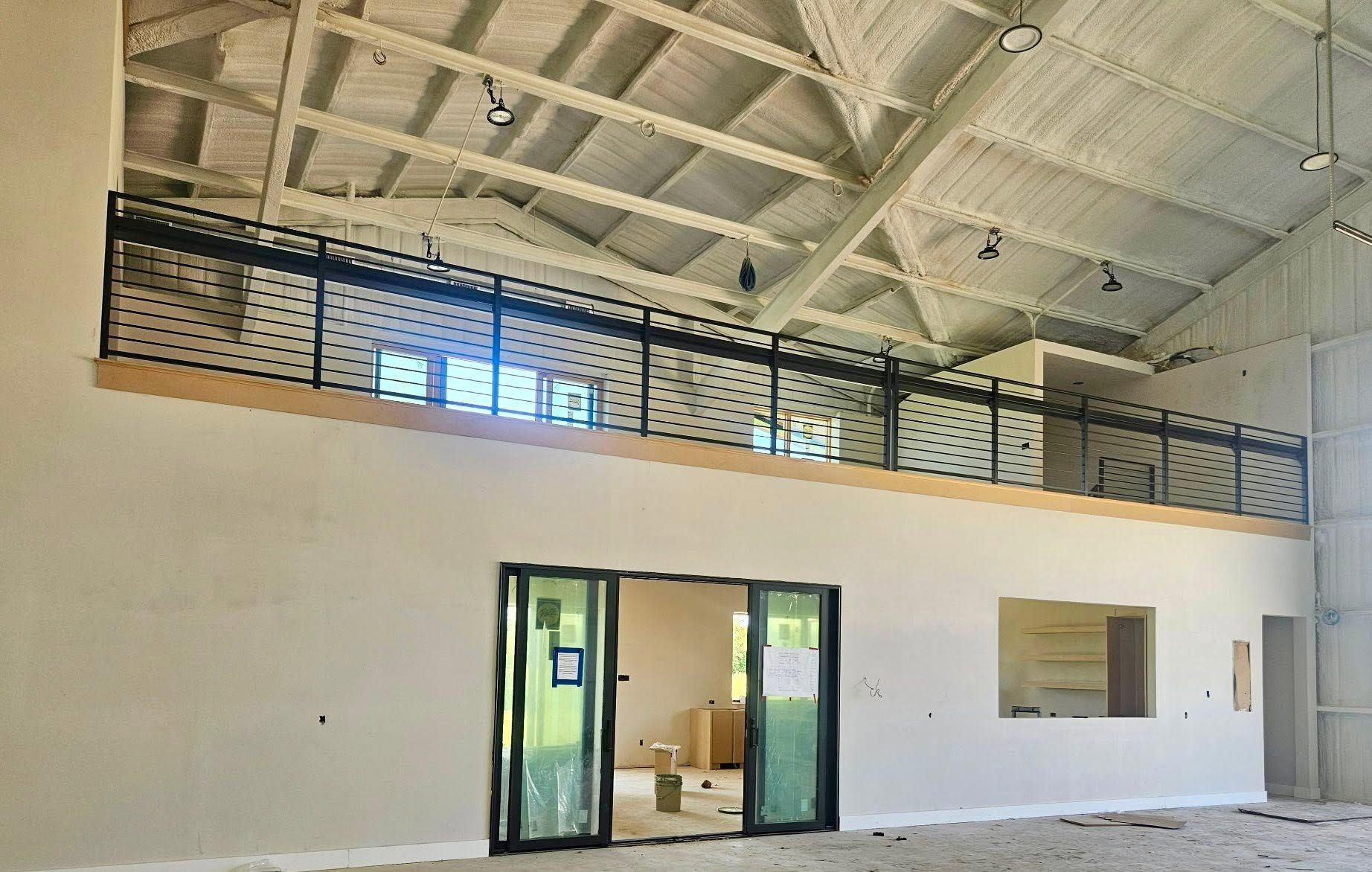 Empty gymnasium with a high ceiling, mezzanine railing, and glass doors along a white wall.