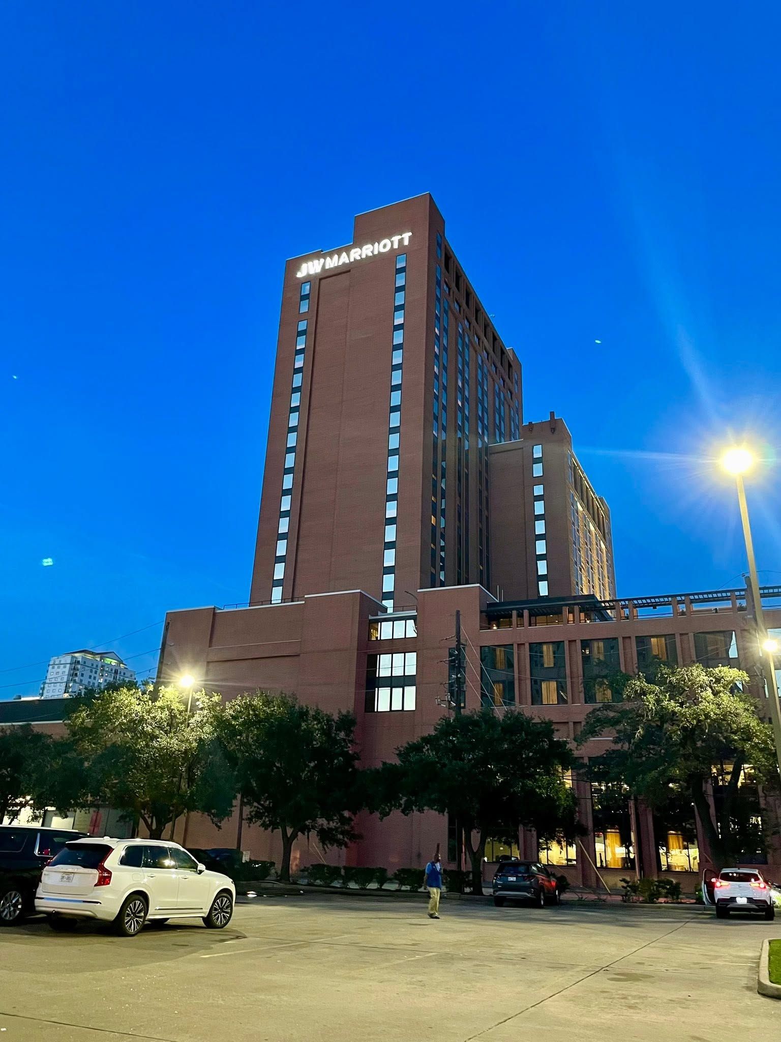 Tall brick high-rise at dusk with parked cars and a bright streetlight in front.
