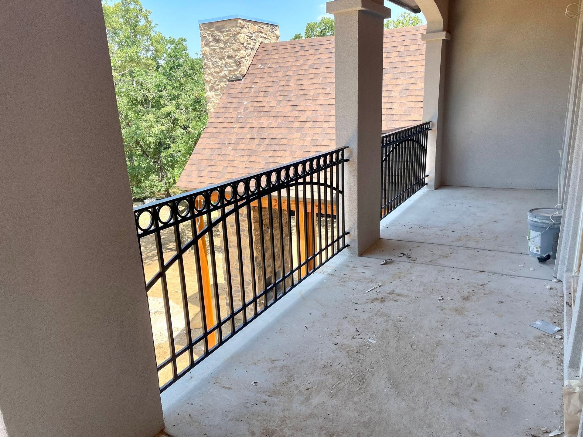 Covered balcony with black railing and beige columns overlooking a tiled roof and trees.