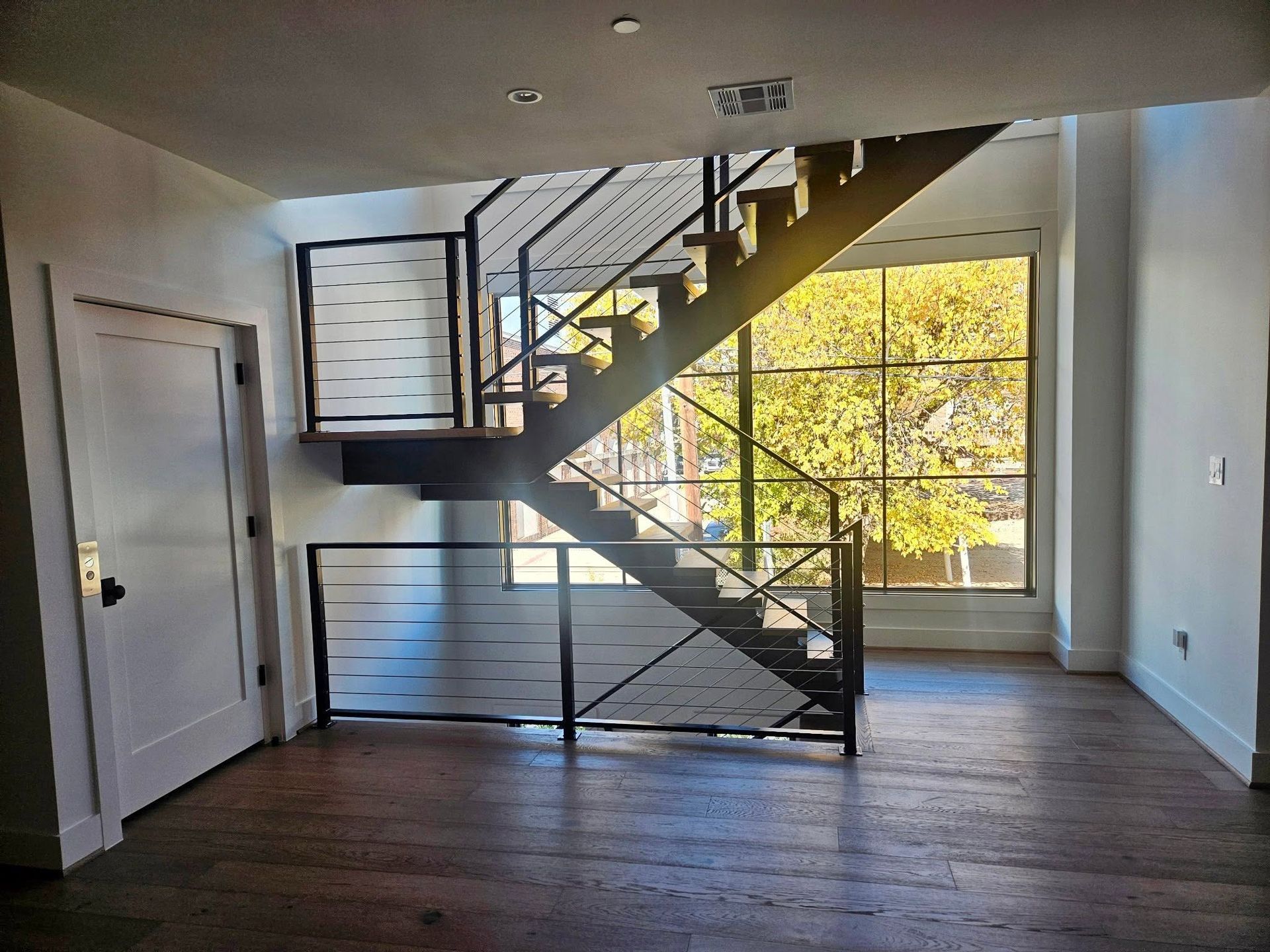 Empty room with stairs and large window, sunlight on wood floor.