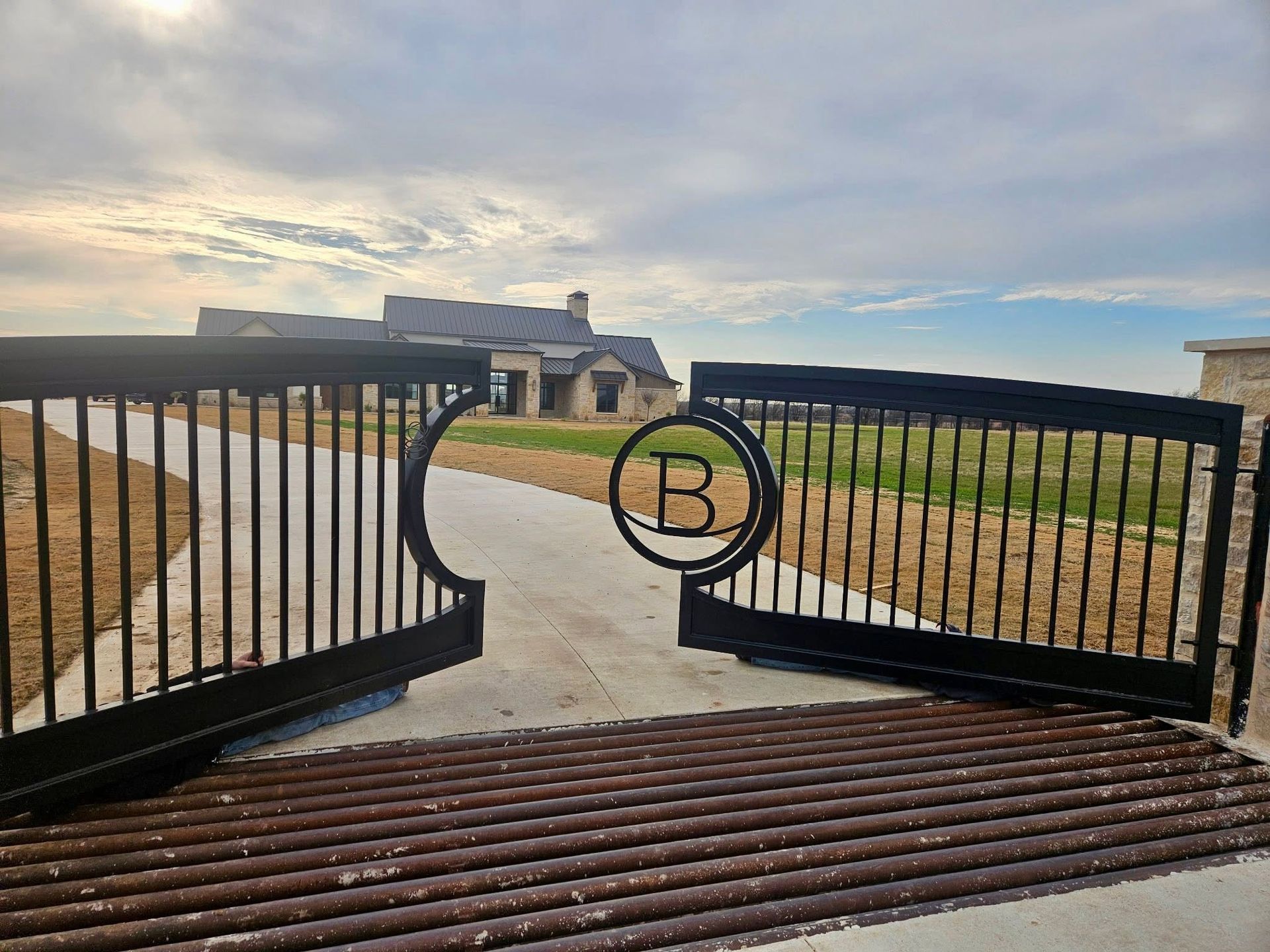 Open black metal gate leading to a paved driveway and house under a cloudy sky.