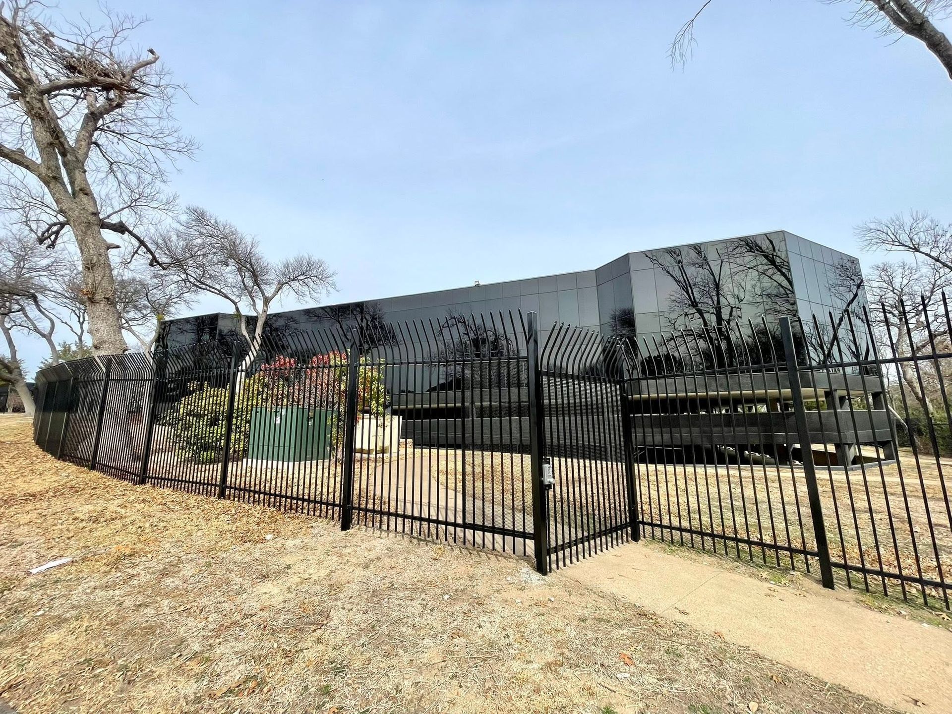 A modern black building behind a black metal fence beside a dry grassy field and bare trees.