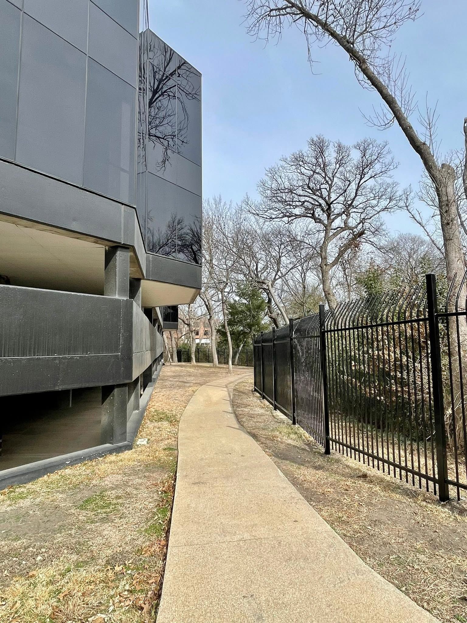 Narrow concrete path beside a gray building and black fence in a park-like setting.