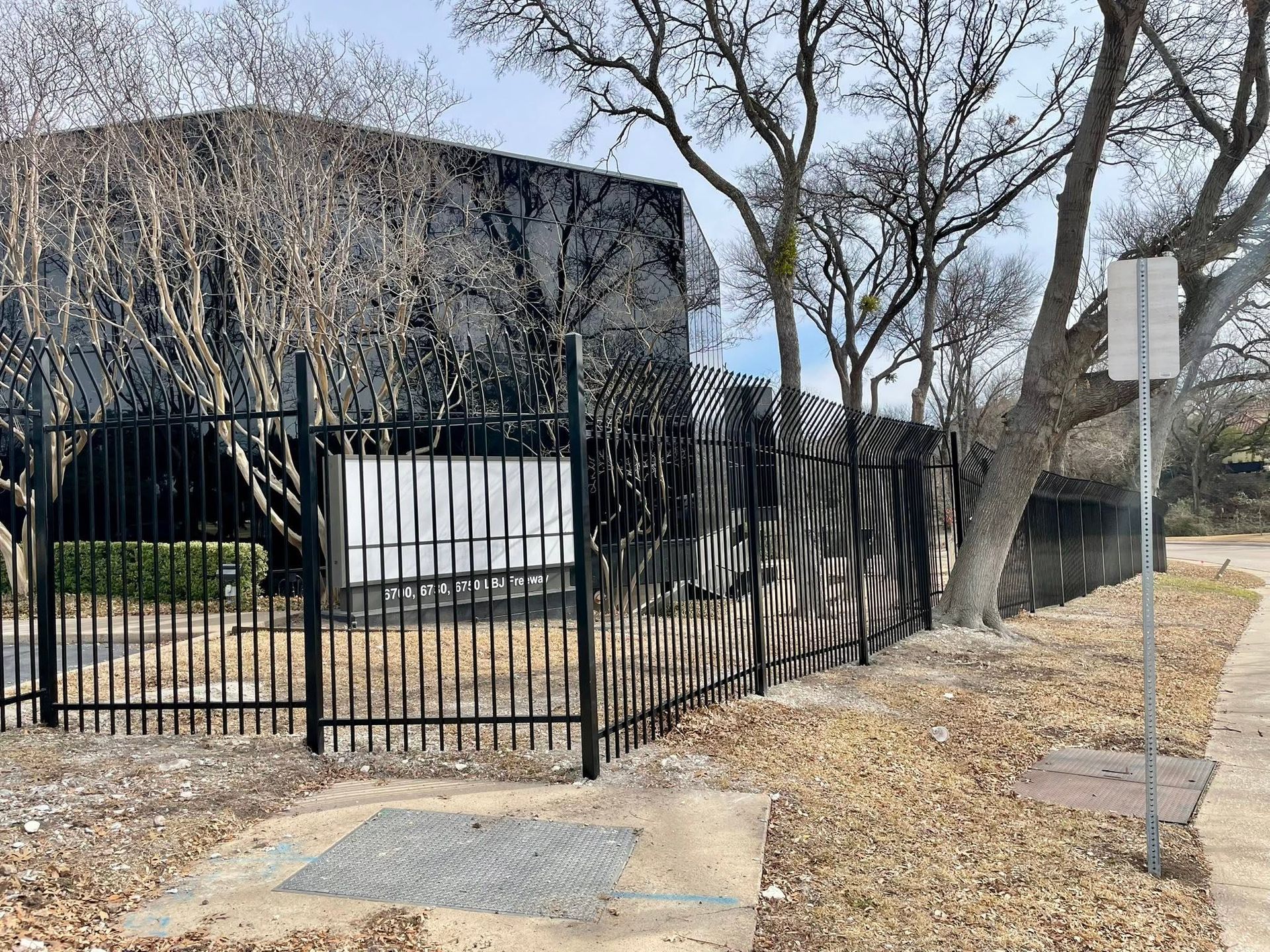 A black metal fence around a dark building beside leafless trees and a sidewalk under a gray sky.