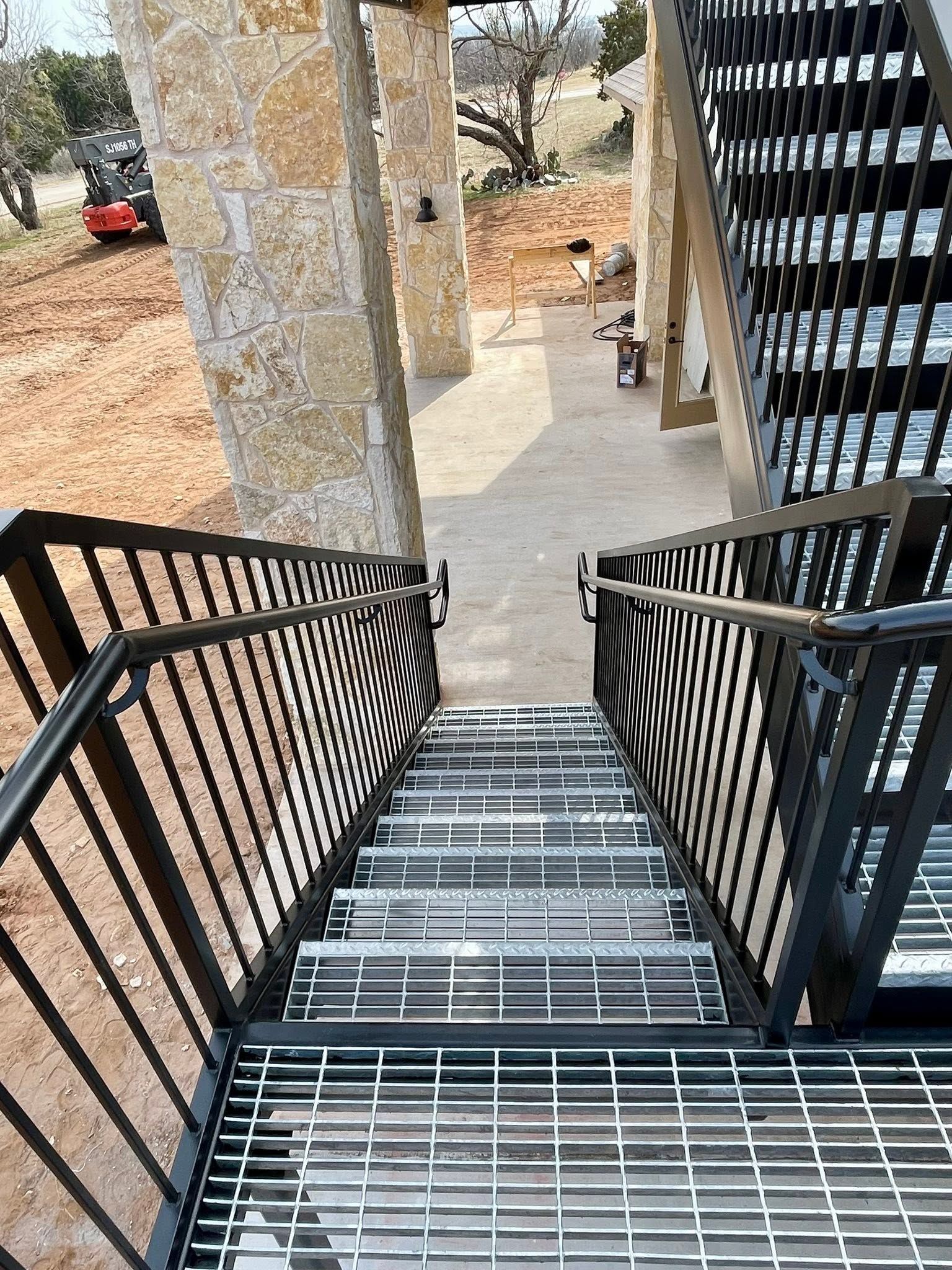 A black metal staircase leading down to a stone walkway beside a building, with railings and a columned porch.