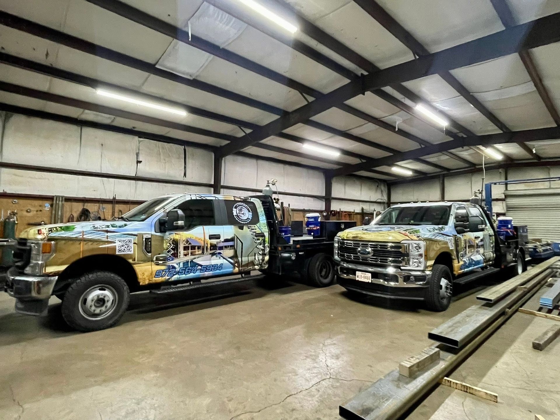 Two modified pickup trucks in a garage, one wrapped in colorful graphics, under bright overhead lights.