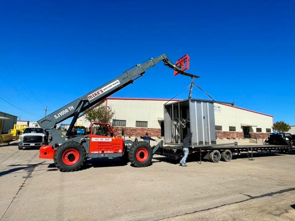 An orange crane is loading a gray structure onto a flatbed truck in an industrial yard.