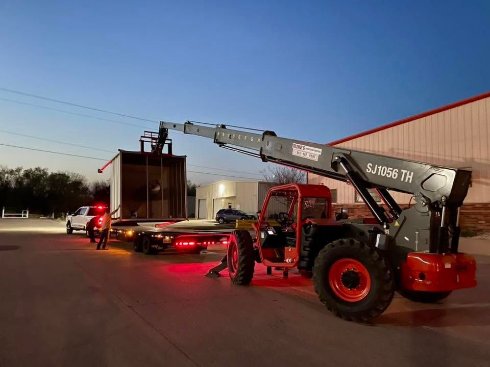 Telehandler lifting a large container near a semi-truck at dusk, with red emergency lights glowing nearby.
