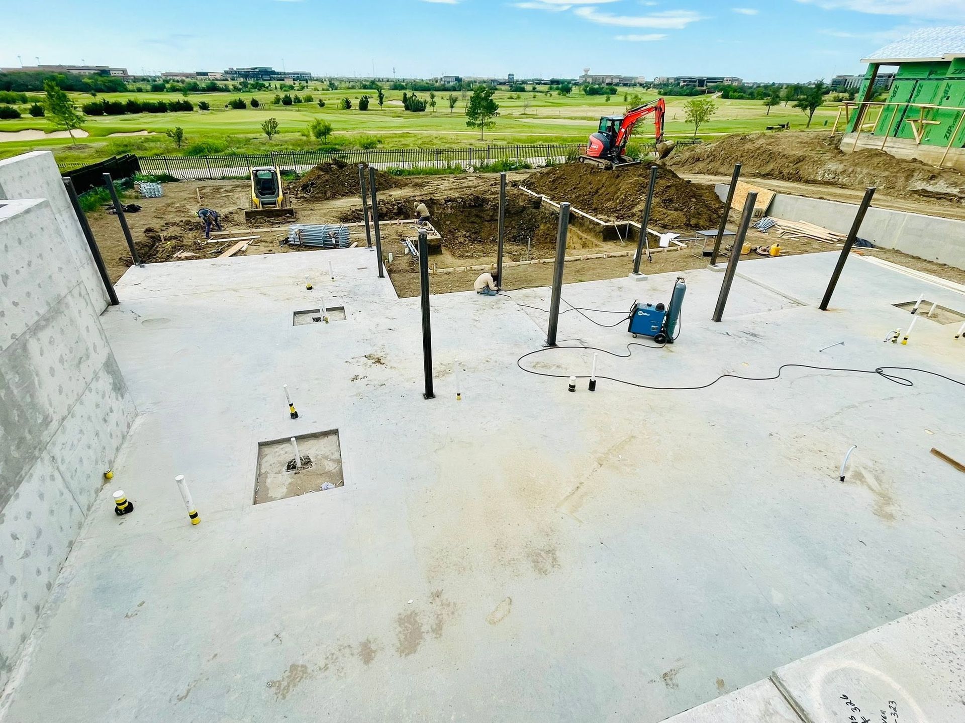Concrete foundation under construction with rebar, overlooking green fields and a few workers in the distance.