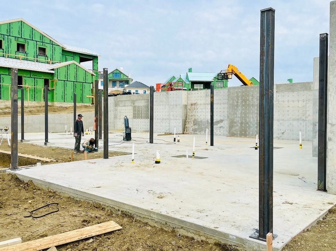 Construction site with concrete foundation slab, steel posts, and workers near unfinished green-framed buildings.