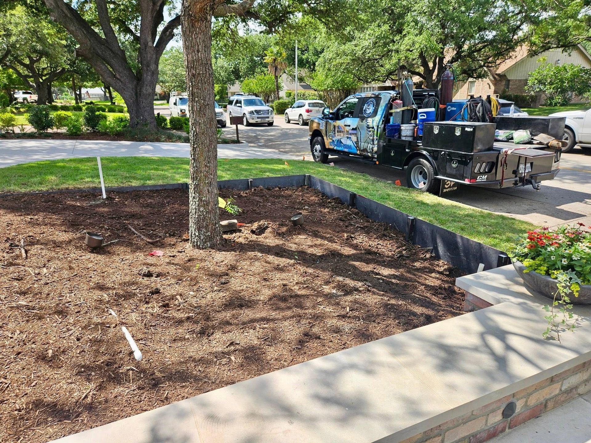 Landscaped yard with mulch bed and young tree, while a work truck and crew are parked nearby.