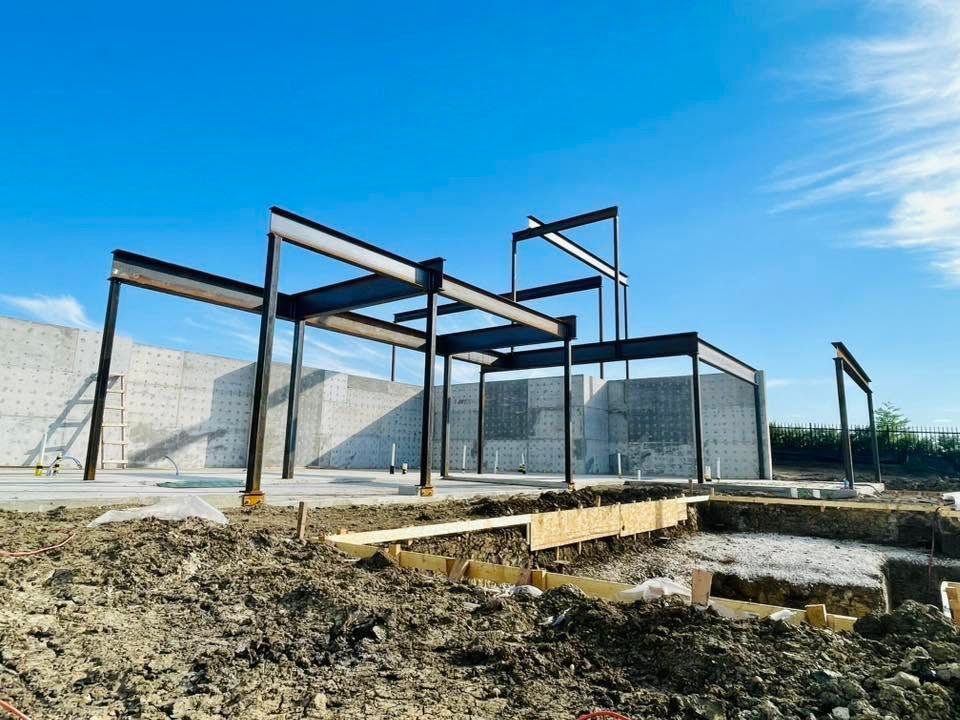 Modern steel-frame building under construction on a dirt lot beneath a blue sky.