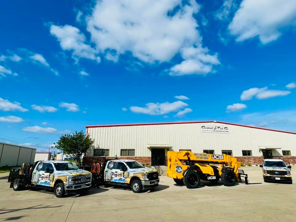 Trucks and heavy machinery are parked outside a beige industrial building under a blue sky.