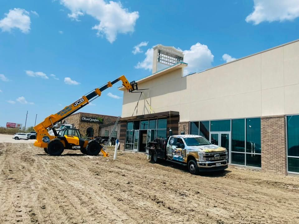 Telehandler lifting materials by a storefront under construction, with a pickup truck parked nearby.