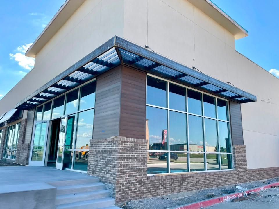 Storefront corner with large glass windows, brick columns, and a metal awning on a sunny day.