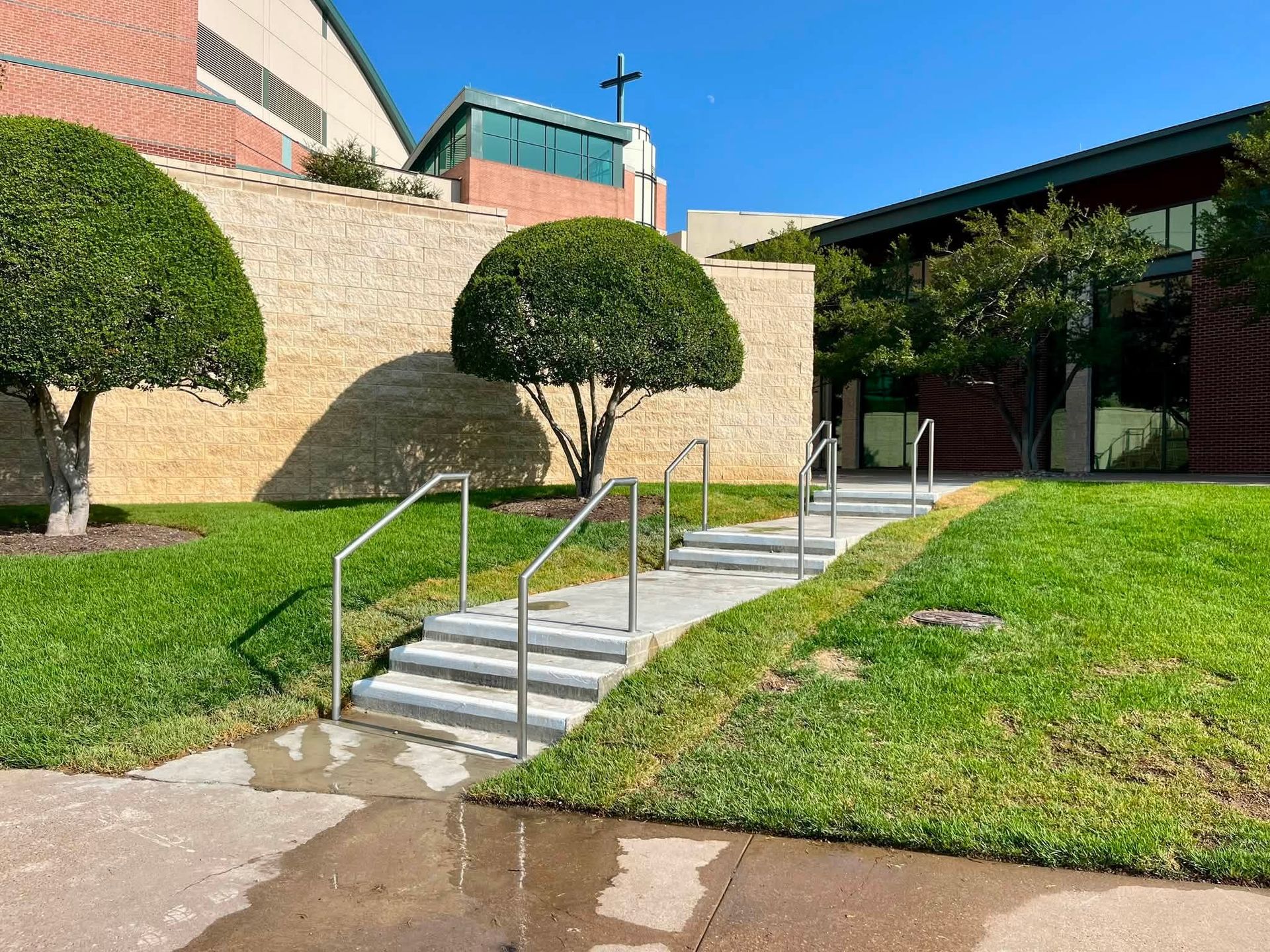 Stone steps with metal railings lead up a grassy hillside beside a brick building and a cross-topped tower.