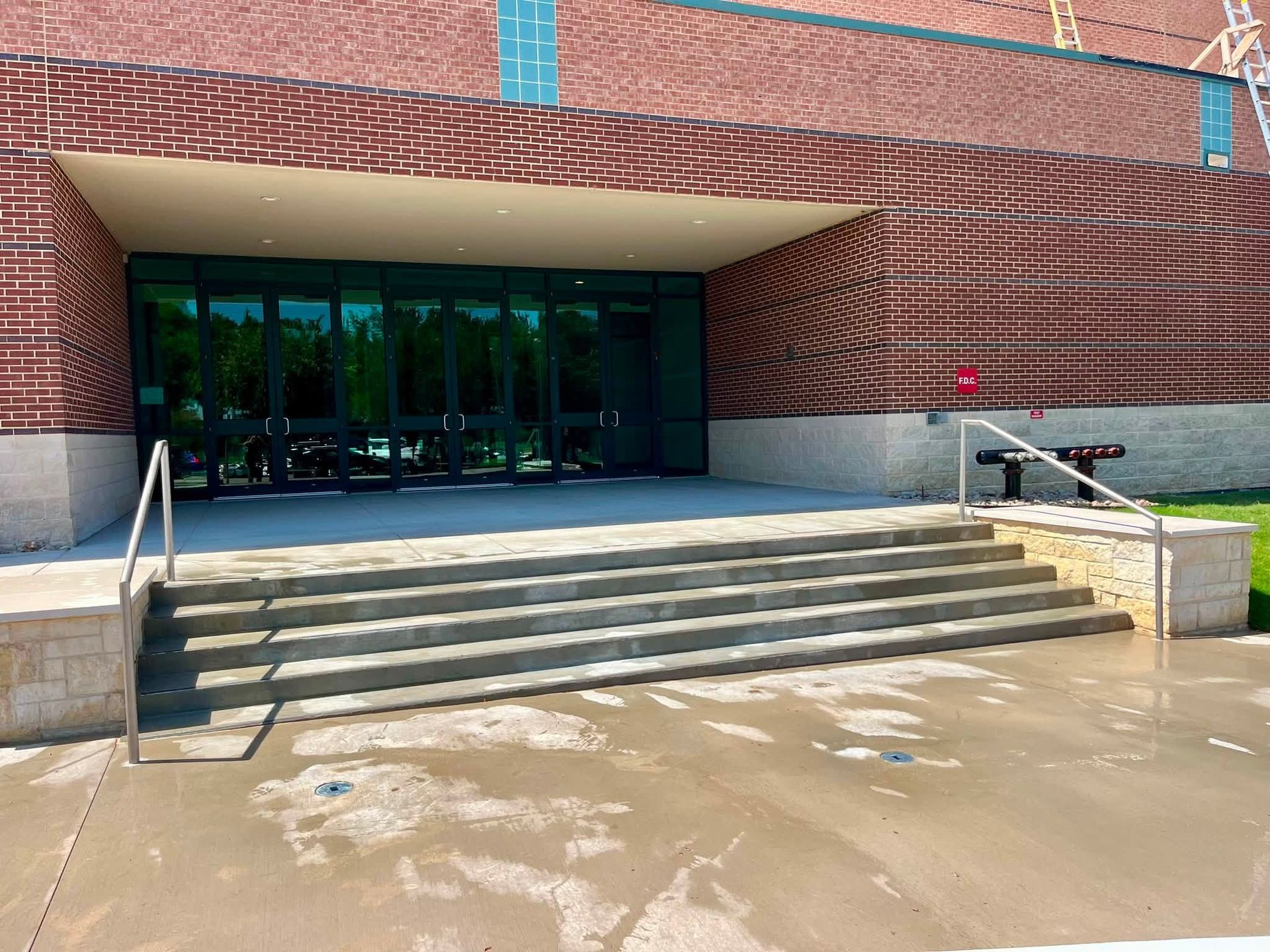 Brick building entrance with steps, handrails, and glass doors reflected in water on the ground.