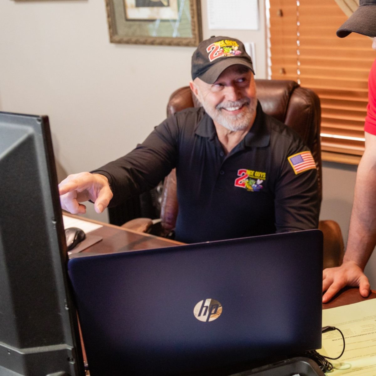 Man in black shirt and cap smiles at a laptop, pointing. A desk, computer screen, and an American flag patch are visible.