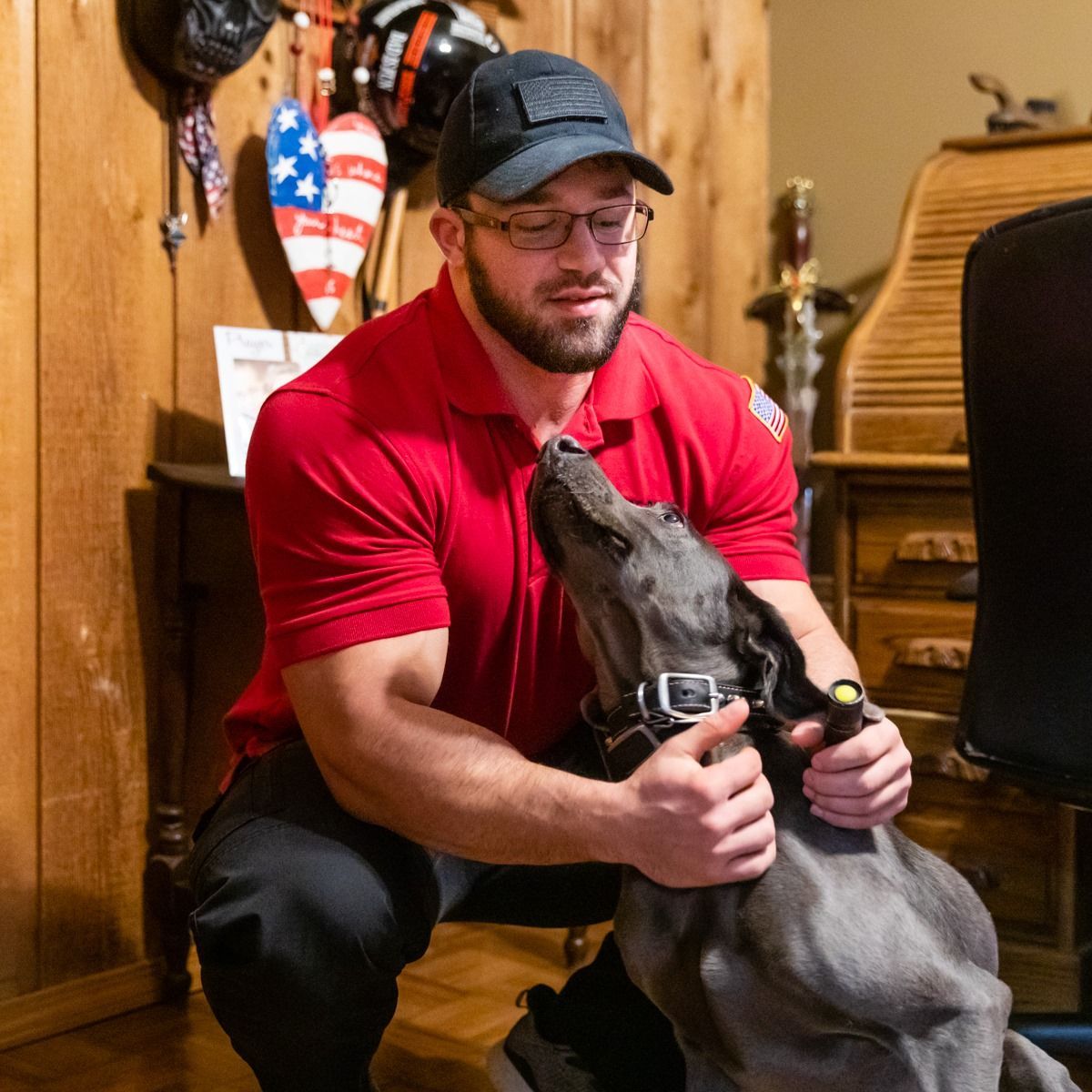 Man in red shirt and glasses petting a gray dog, wooden interior.