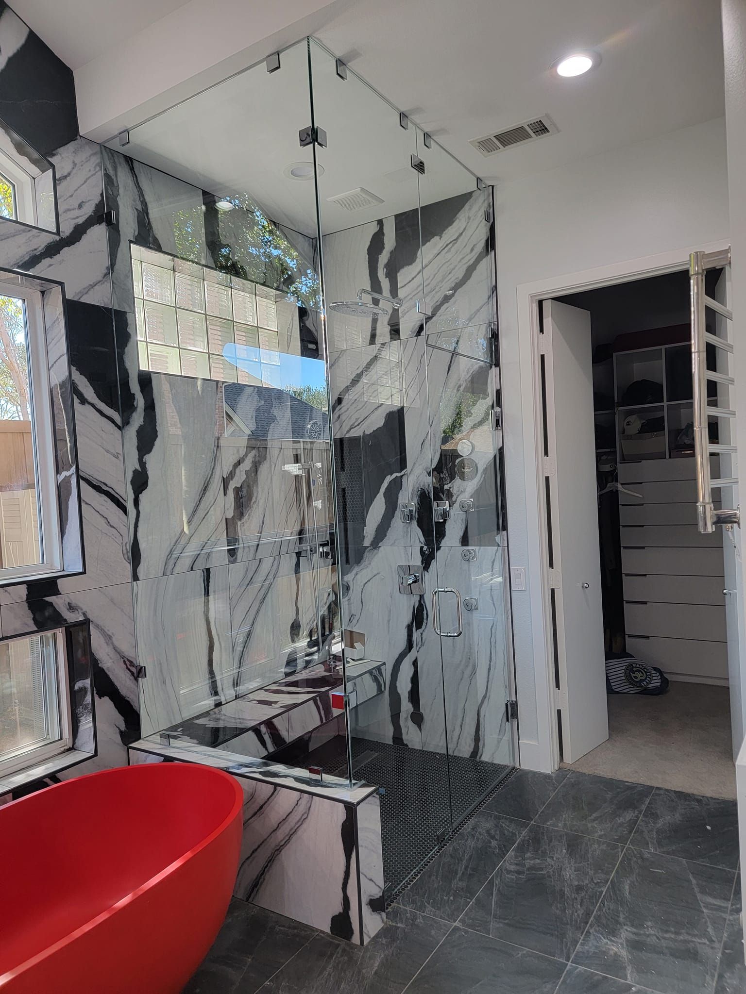 Modern bathroom with marble-pattern walls, glass shower partition, red tub, and open doorway to a bedroom