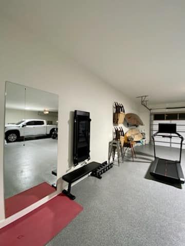 Small gym room with mirror, exercise machine, treadmill, and a parked white car visible through the open doorway