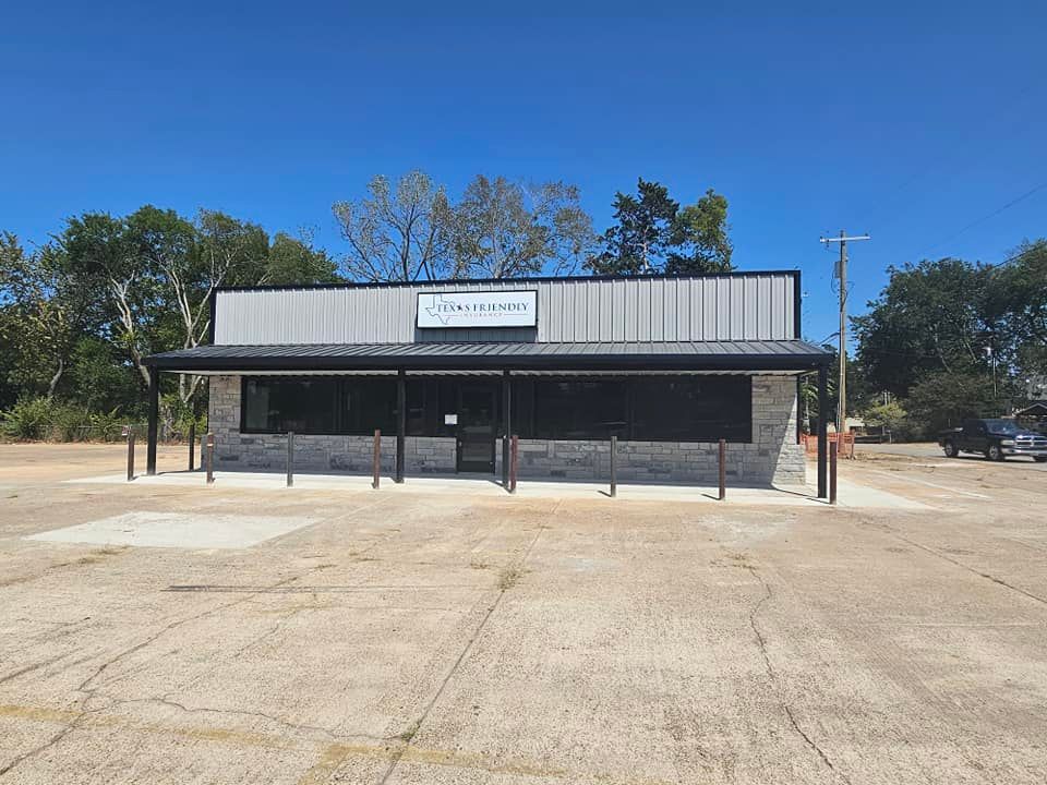 Small roadside building with a metal awning and closed storefront, set in a mostly empty parking lot under a clear sky