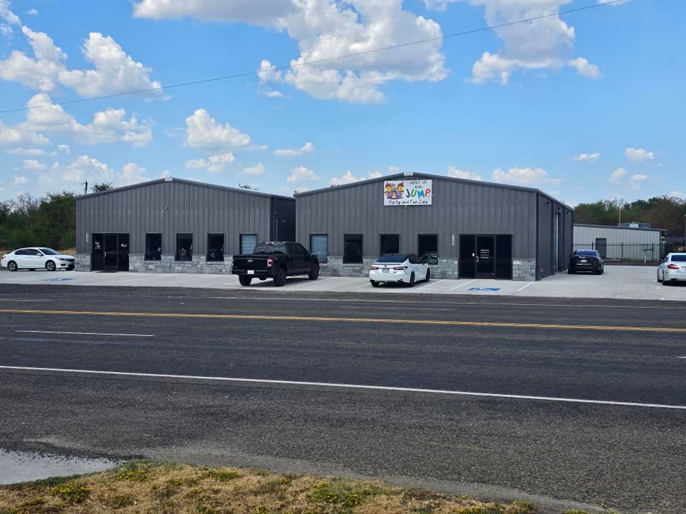 Gray industrial building with parked cars under a partly cloudy blue sky.