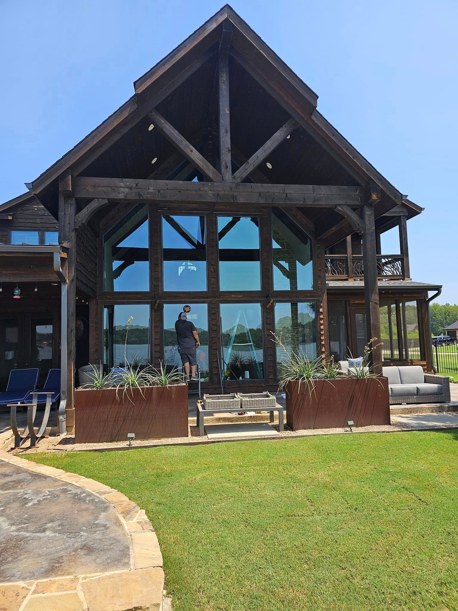 Glass-front lodge with dark wood beams, a grassy lawn, and patio seating under a blue sky