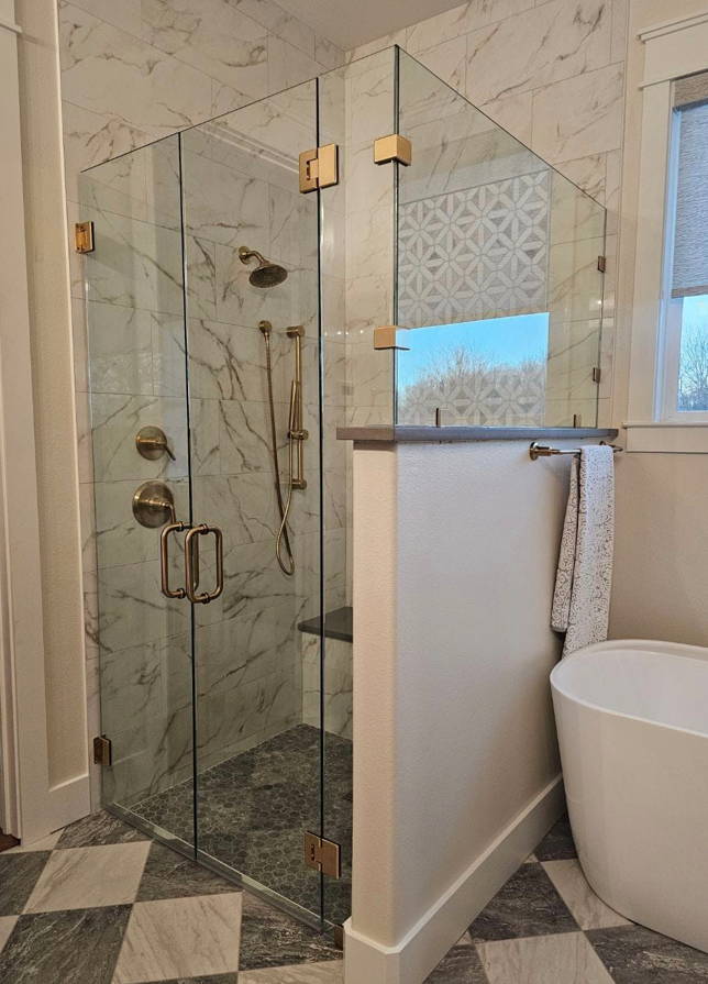 Glass shower enclosure beside a tiled bathtub area with a window and towel rack