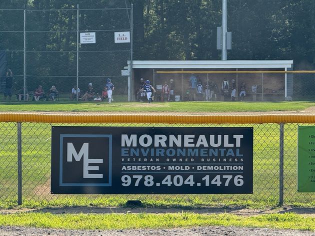 A banner for Morneault Environmental on a chain-link fence at a baseball field with players in the background.