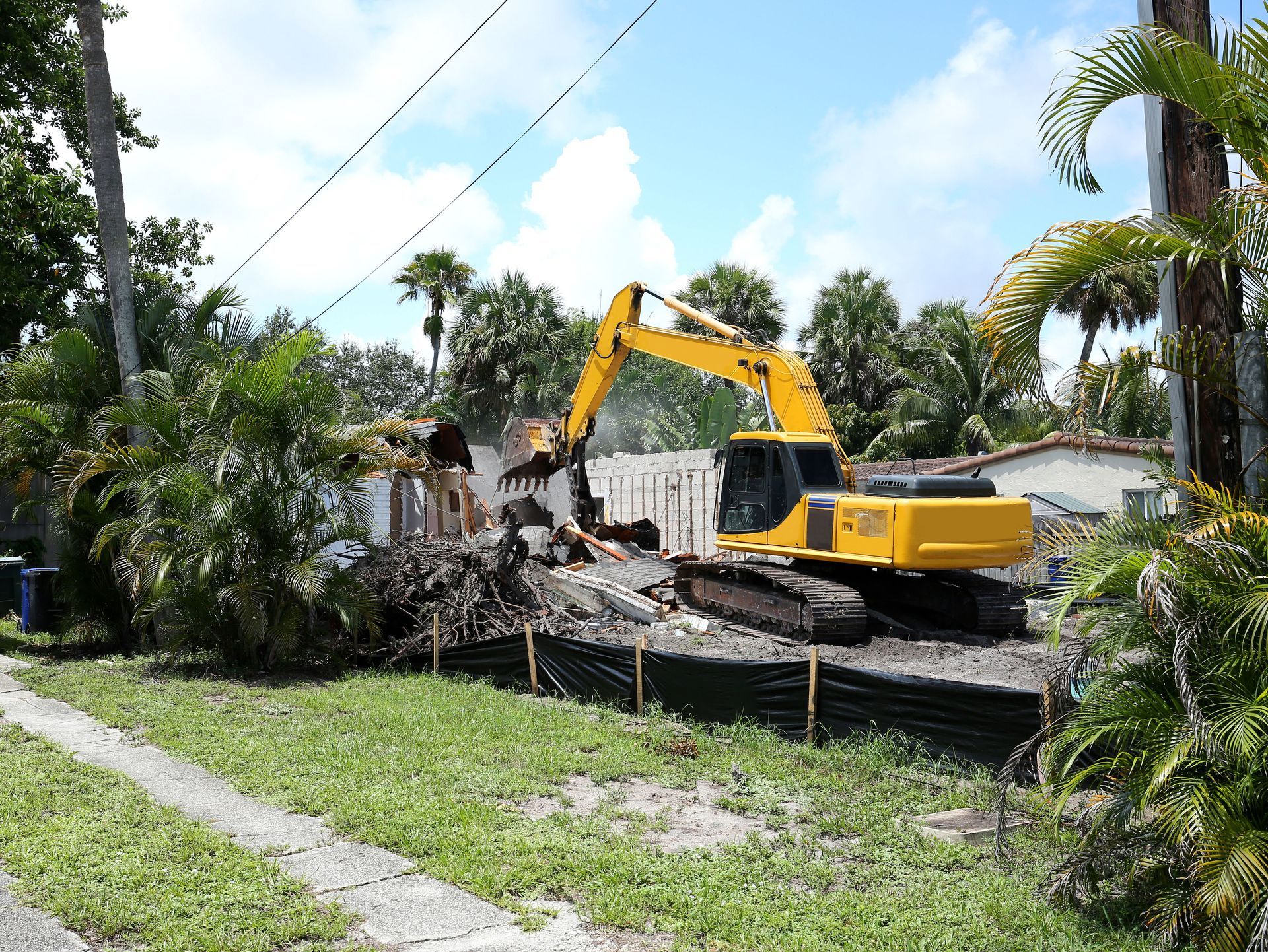 A yellow excavator tears down a residential structure behind a black mesh fence in a sunny, tree-filled neighborhood.