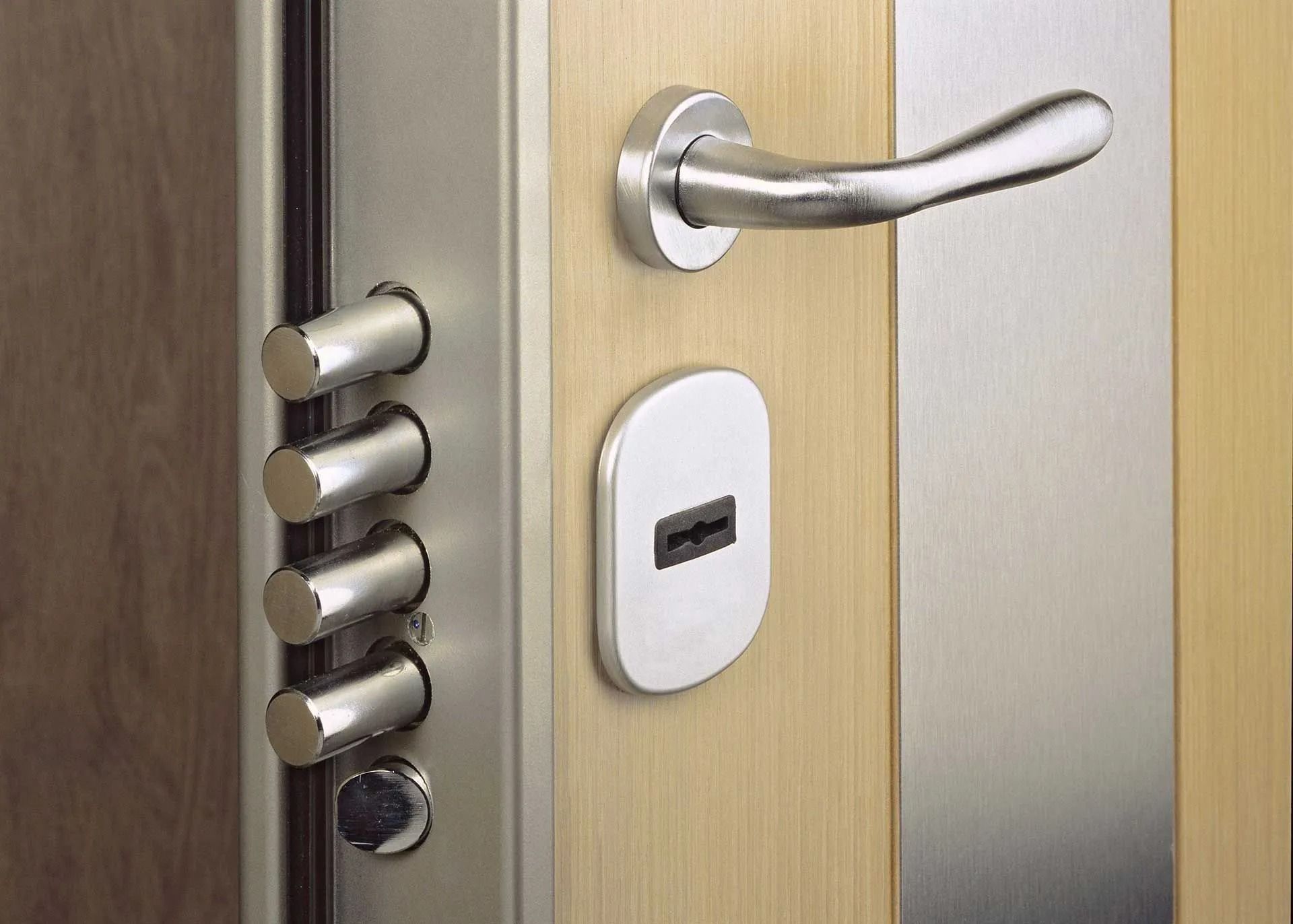 Close-up of a door with multiple locking bolts, a handle, and a keyhole, all in silver against a light-colored wood door.
