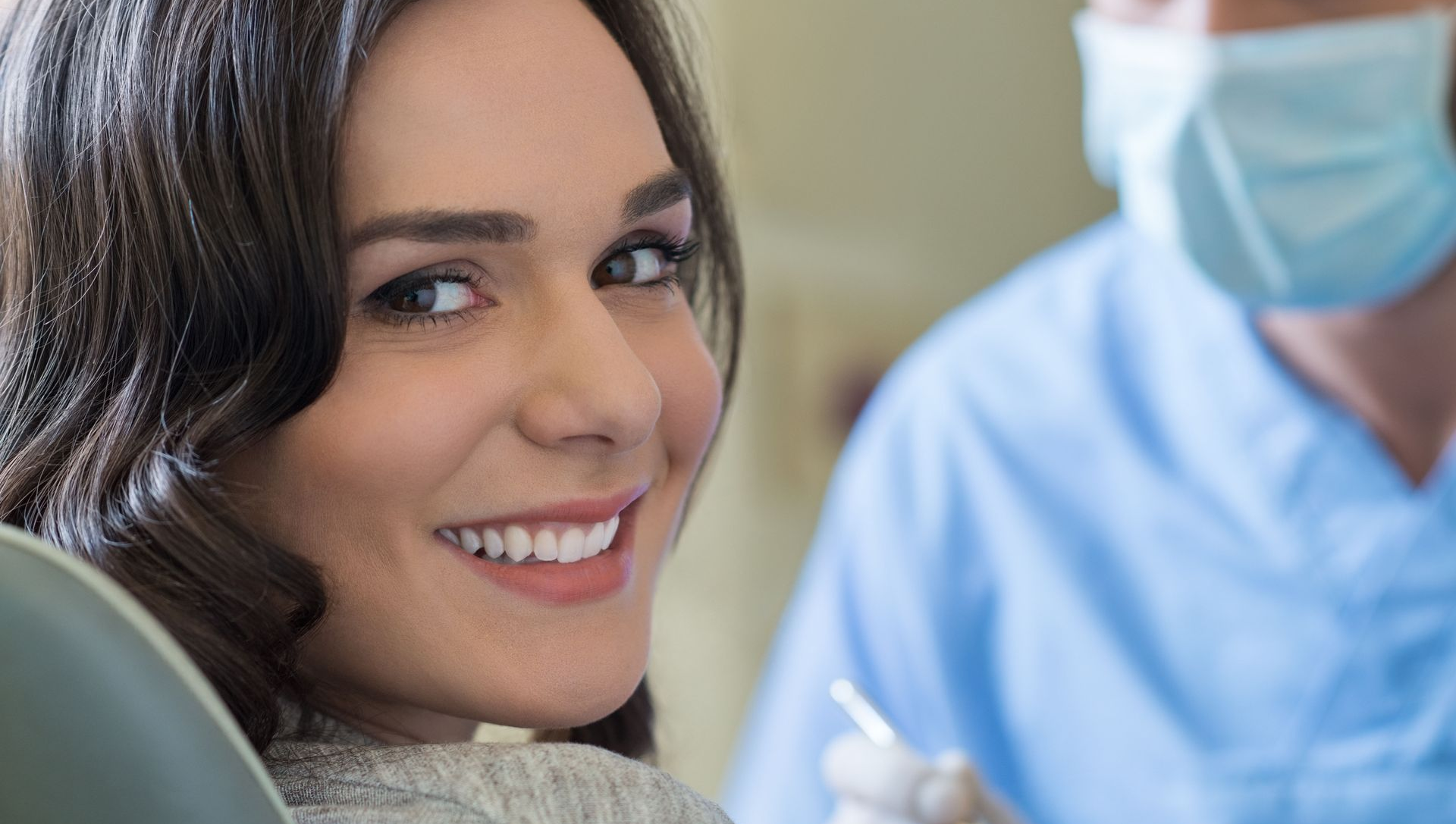 A smiling patient in a dental chair looks back at a healthcare professional in a mask and blue scrubs.