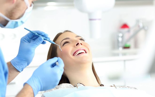 A dentist in blue gloves uses a dental tool to examine the teeth of a smiling patient in a bright, modern office.