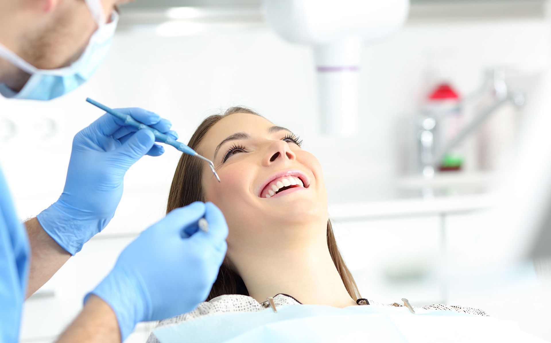 A dentist in blue gloves uses a dental tool to examine the teeth of a smiling patient in a bright, modern office.