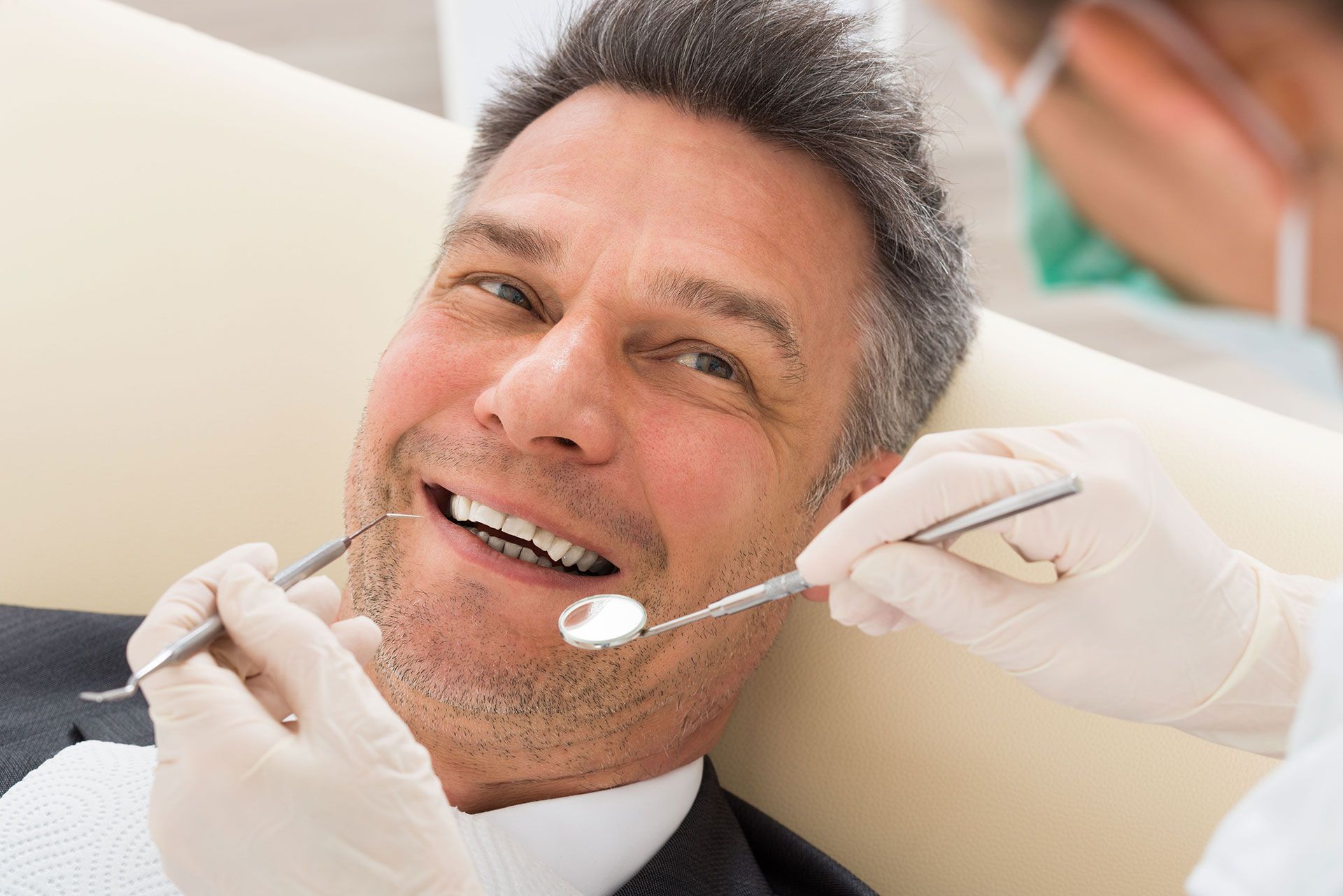 A dentist wearing white gloves uses a mirror and dental tool to examine a patient's teeth in a clinical setting.