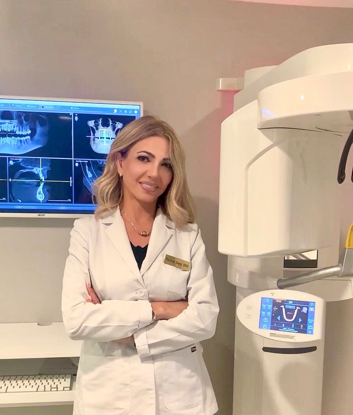 Woman in white coat smiles, arms crossed, beside dental x-ray machine in a clinic setting.