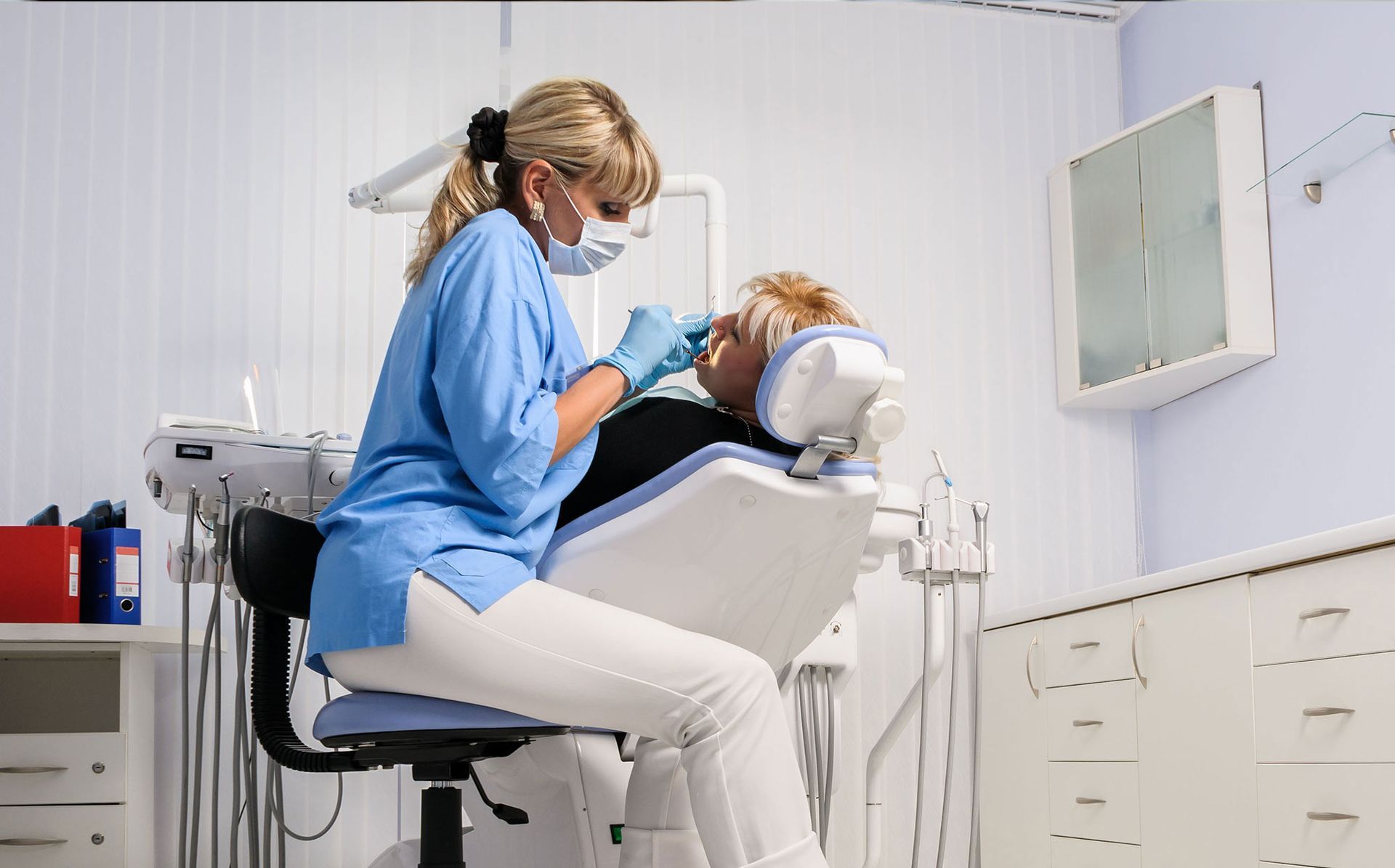 A dental professional in blue scrubs and a face mask performing an examination on a patient in a dental chair.