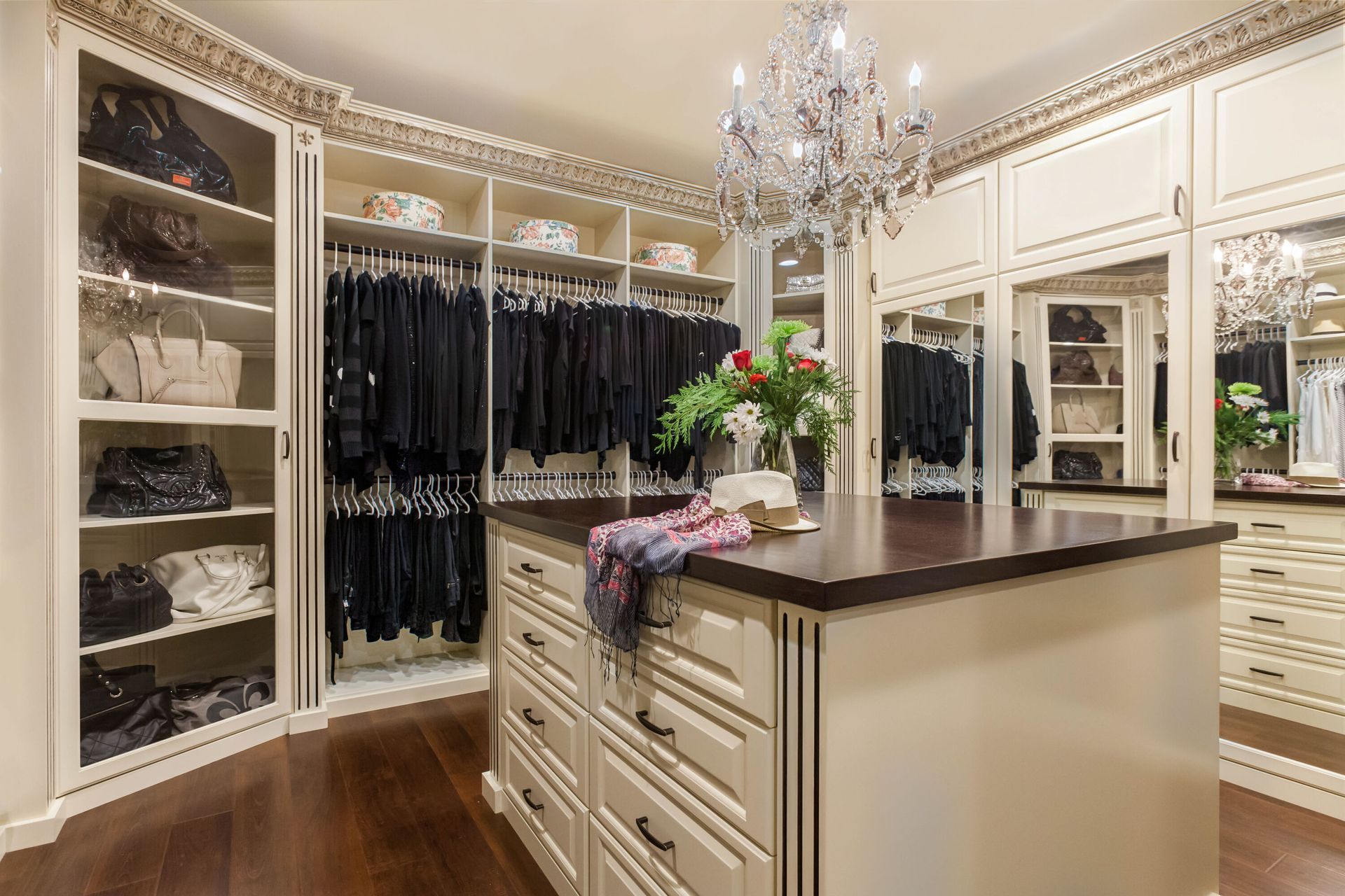 Elegant walk-in closet with cream cabinetry, dark wood floors, and a central island. Features include a chandelier, clothing racks, and purses.