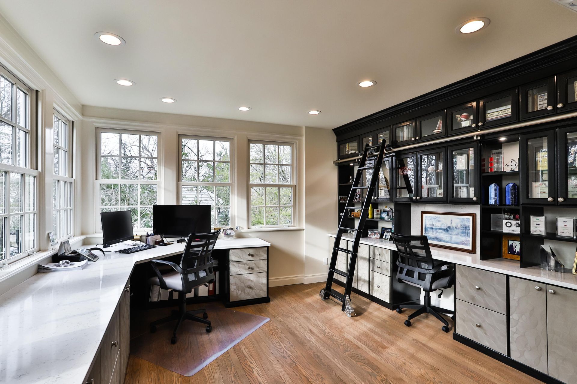 A home office with a wrap-around desk by windows and a built-in black bookcase with a rolling ladder. Two black desk chairs and a wood floor.