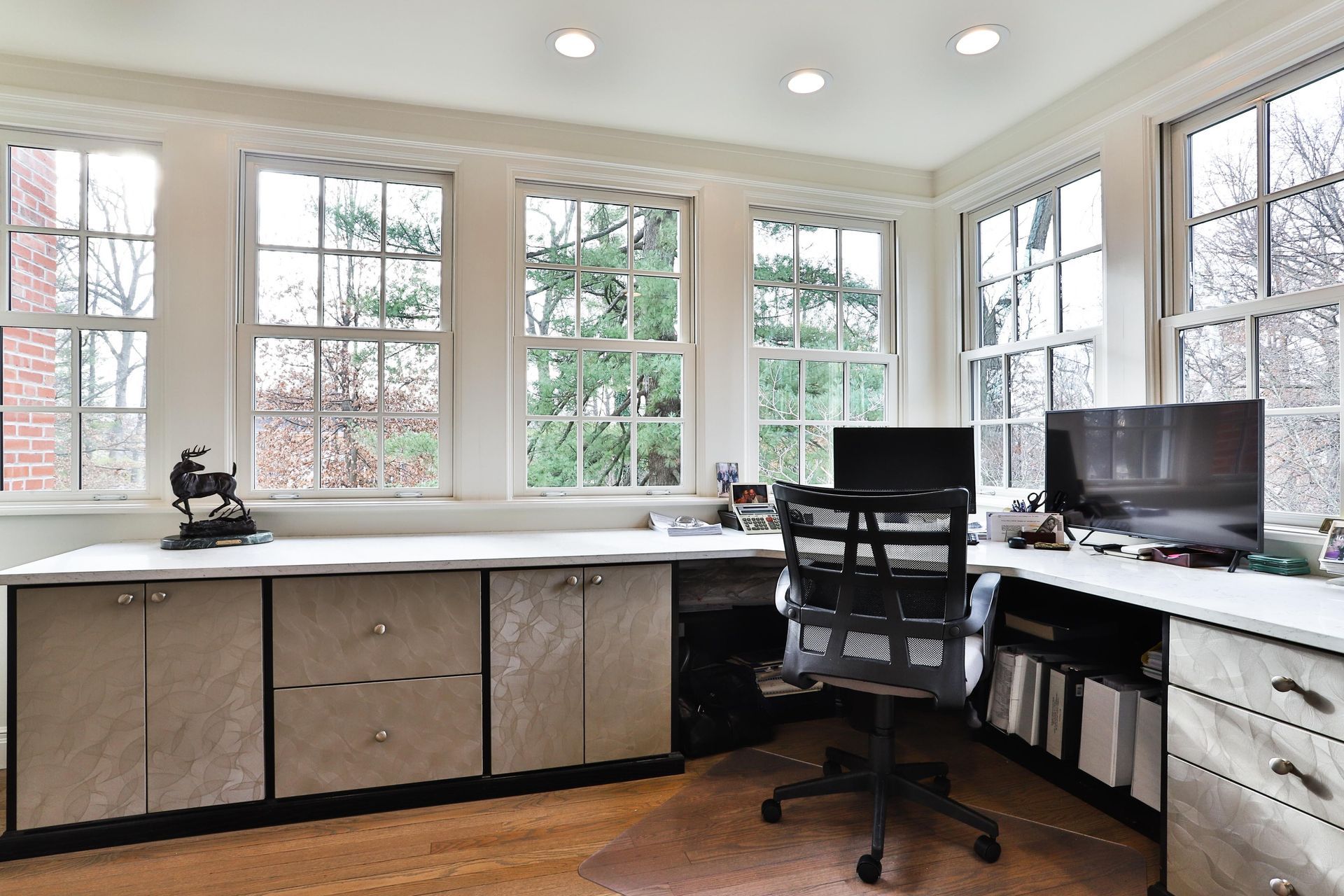 Home office with built-in desk and large windows overlooking trees. A black office chair sits at the desk, with a computer monitor.