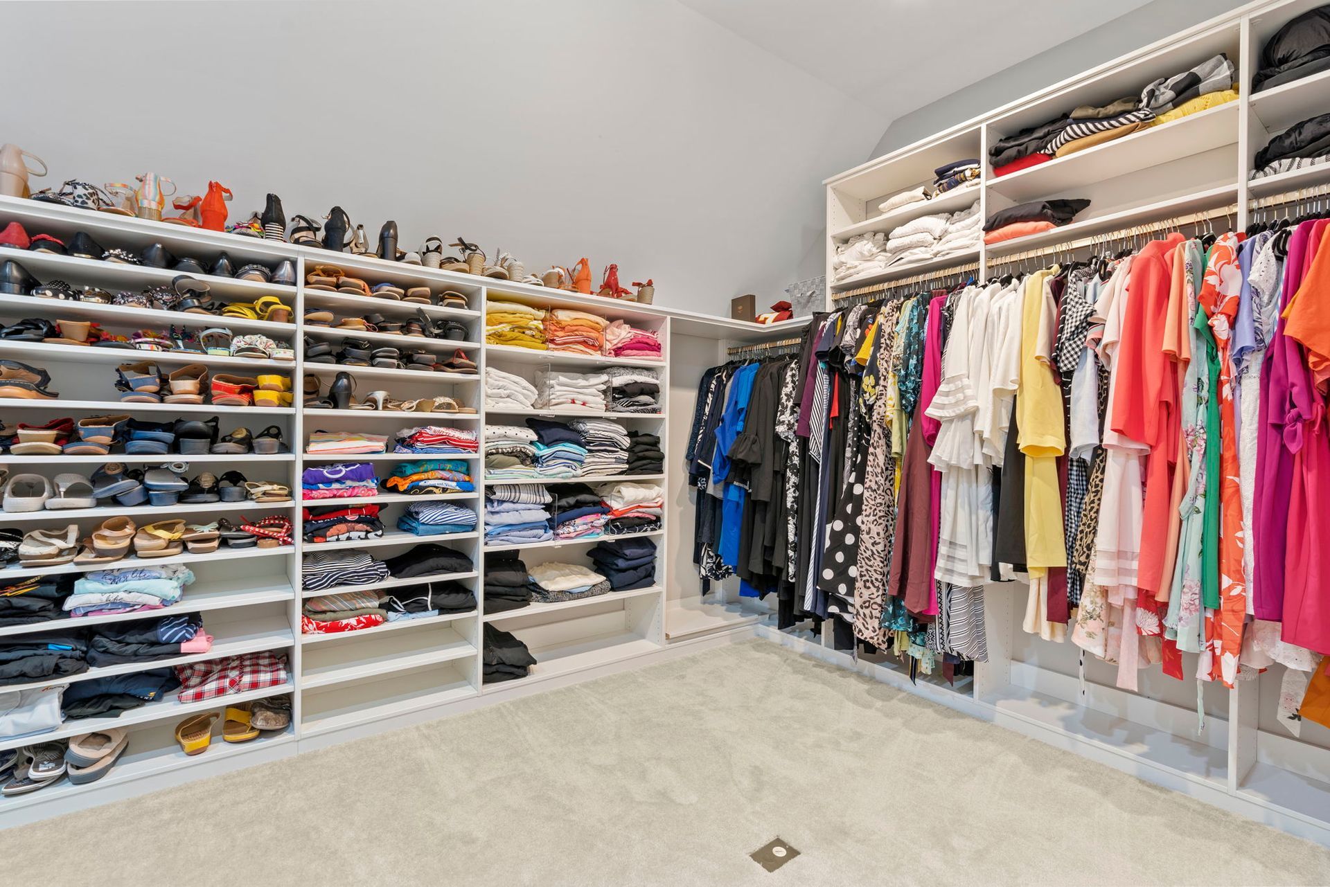 Walk-in closet with white shelving filled with shoes and clothes of various colors. Bright and spacious interior.