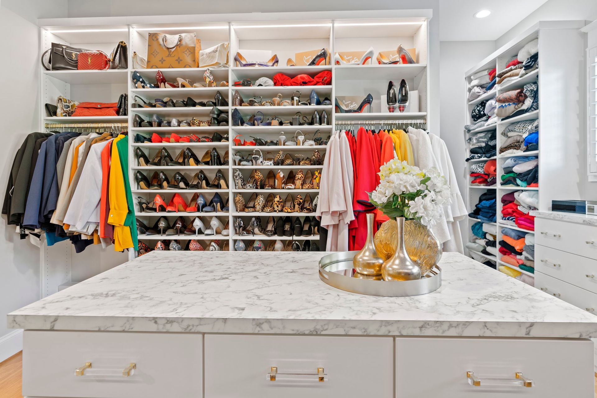 A luxurious walk-in closet with a marble-topped island, shelves of shoes, hanging clothes in various colors, and handbags.