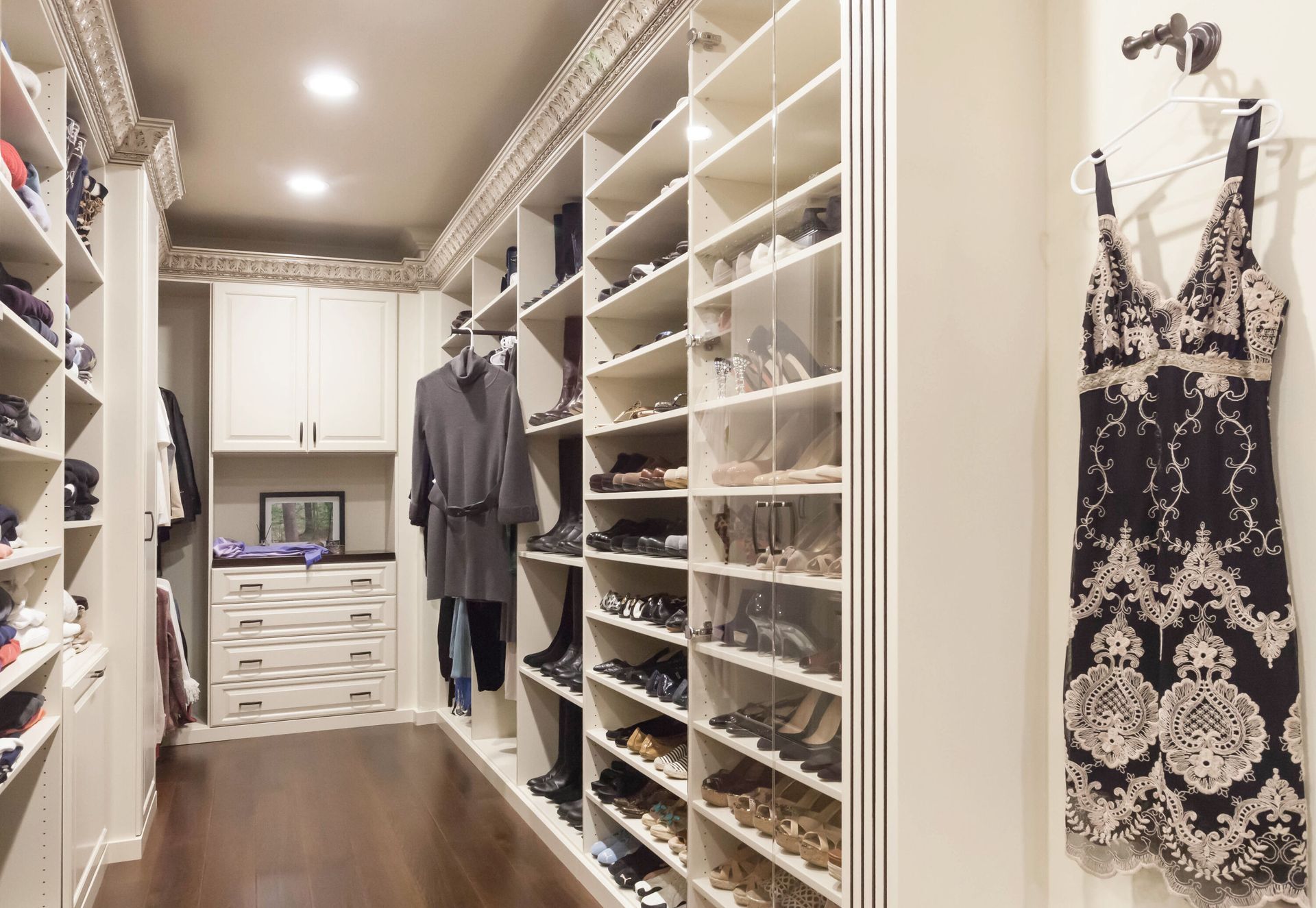 A walk-in closet with cream shelving, packed with shoes and clothes. A dress hangs on the right side.