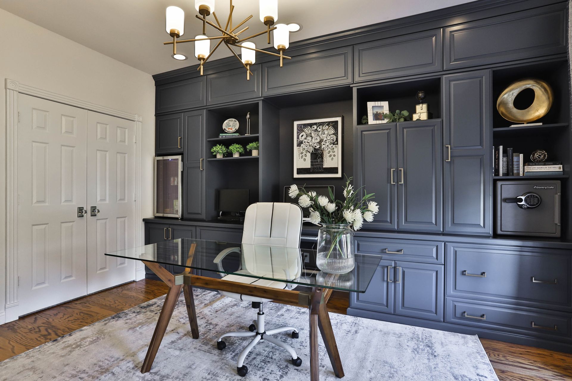 Home office with a dark blue built-in bookcase, glass desk, and white rug. White flowers and gold accents provide a contrast.