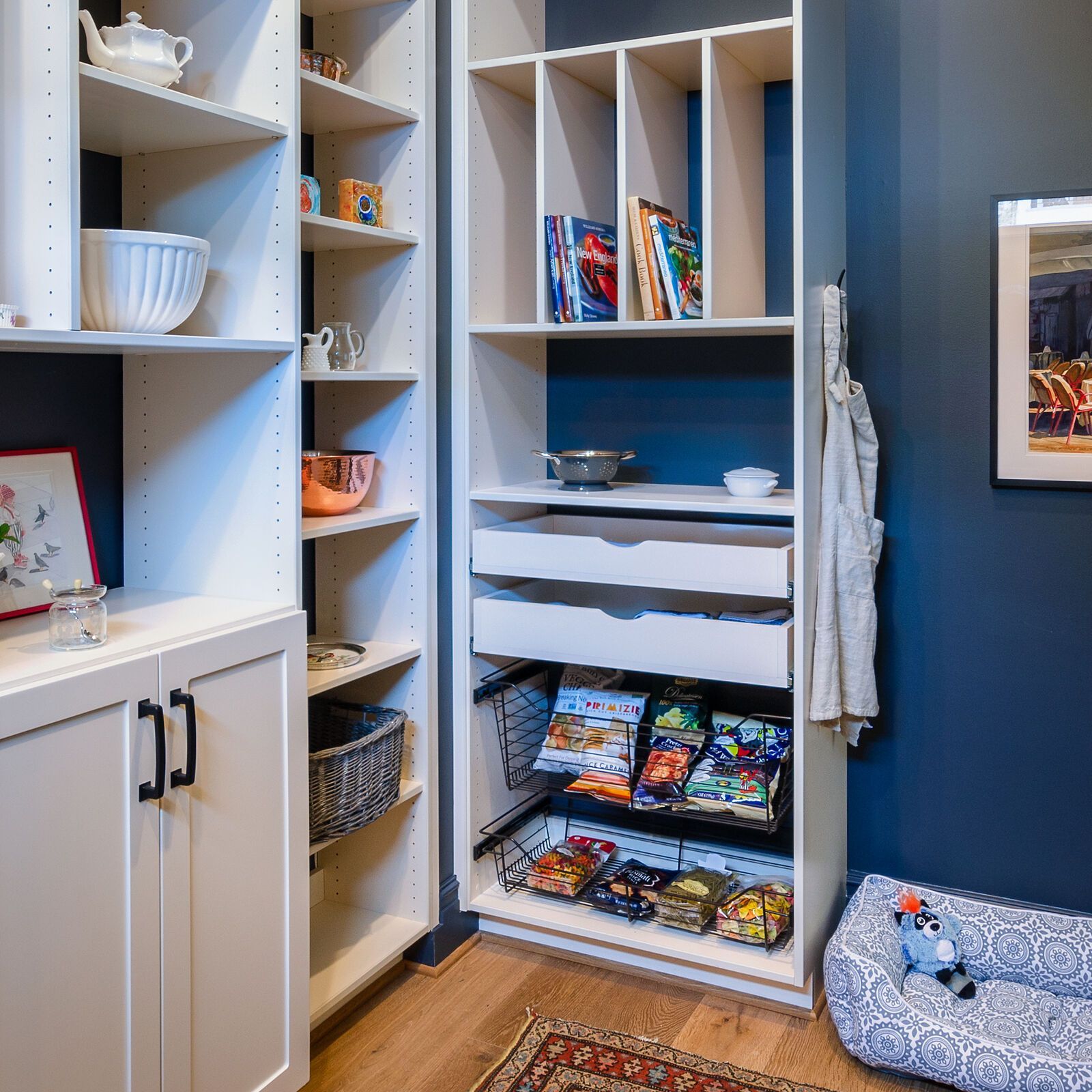 Built-in white shelving unit with storage for books, magazines, and kitchenware, against a dark blue wall, with a dog bed in the corner.