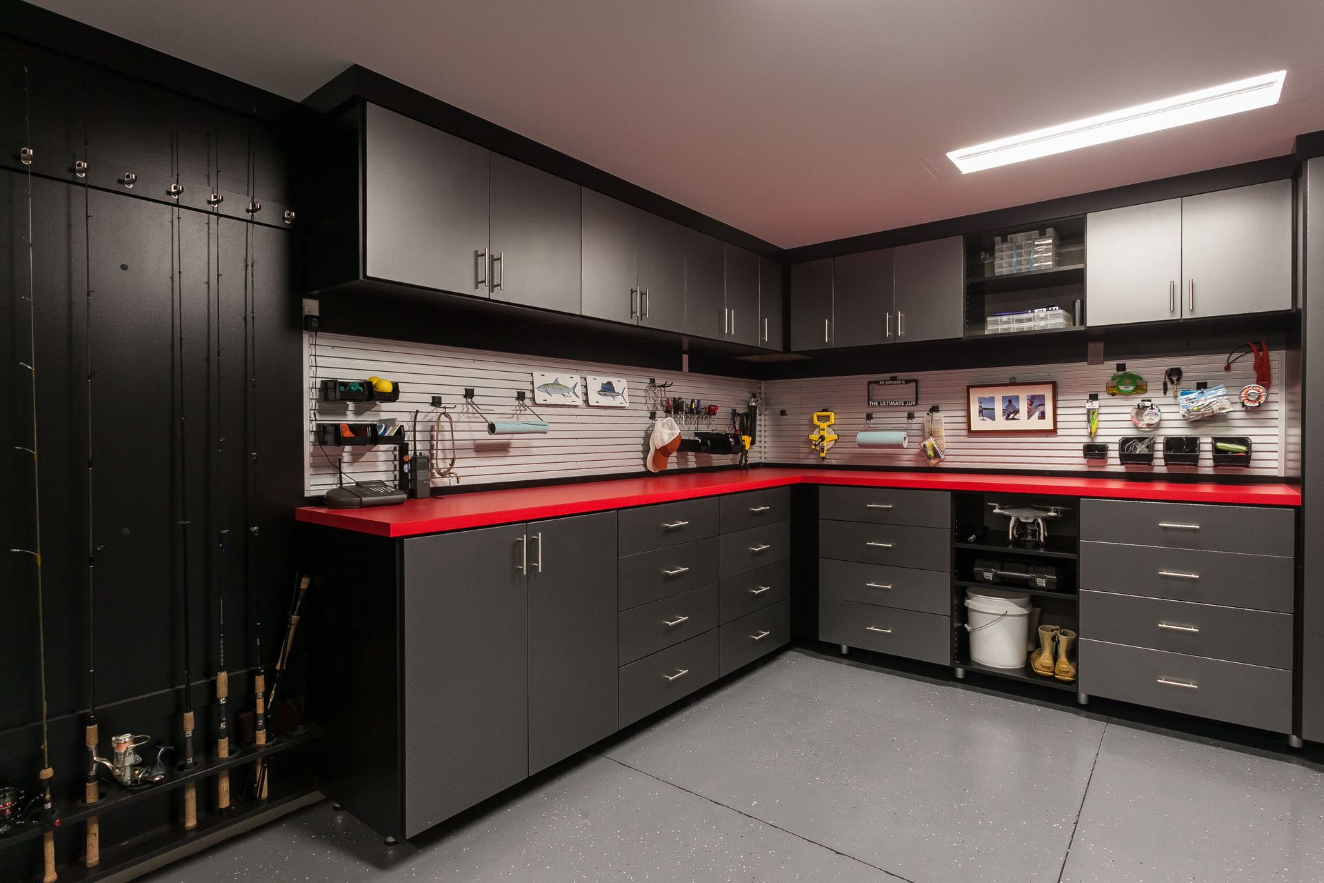 A modern garage with black cabinets, red countertops, and pegboards displaying tools. Fishing rods line one wall.